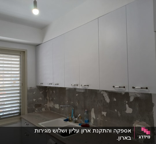 מטבח עם ארונות לבנים, כיור וברז, קיר מתקלף